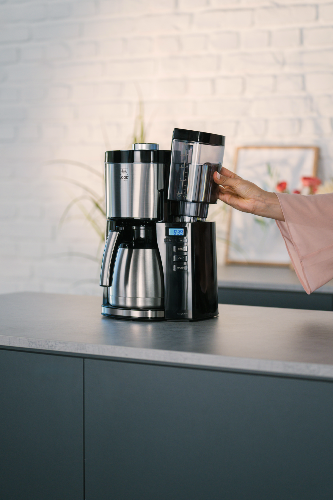 Melitta® LOOK® Therm Timer Filterkaffeemaschine mit Isolierkanne auf einer Küchenarbeitsplatte, während eine Hand den Wassertankdeckel öffnet.