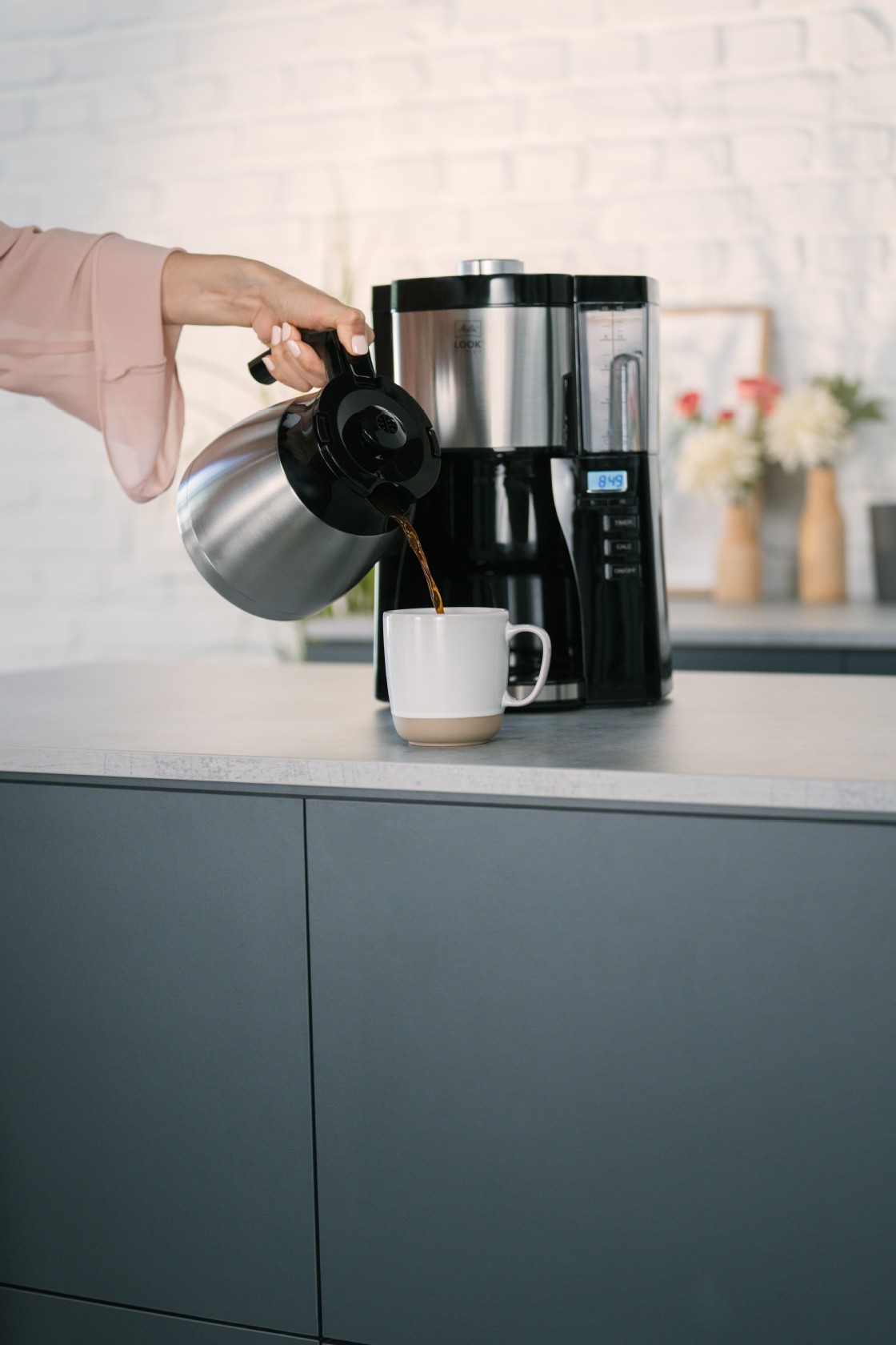 Melitta® LOOK® Therm Timer Filterkaffeemaschine auf einer Küchenarbeitsplatte, während Kaffee aus der Isolierkanne in eine Tasse gegossen wird.