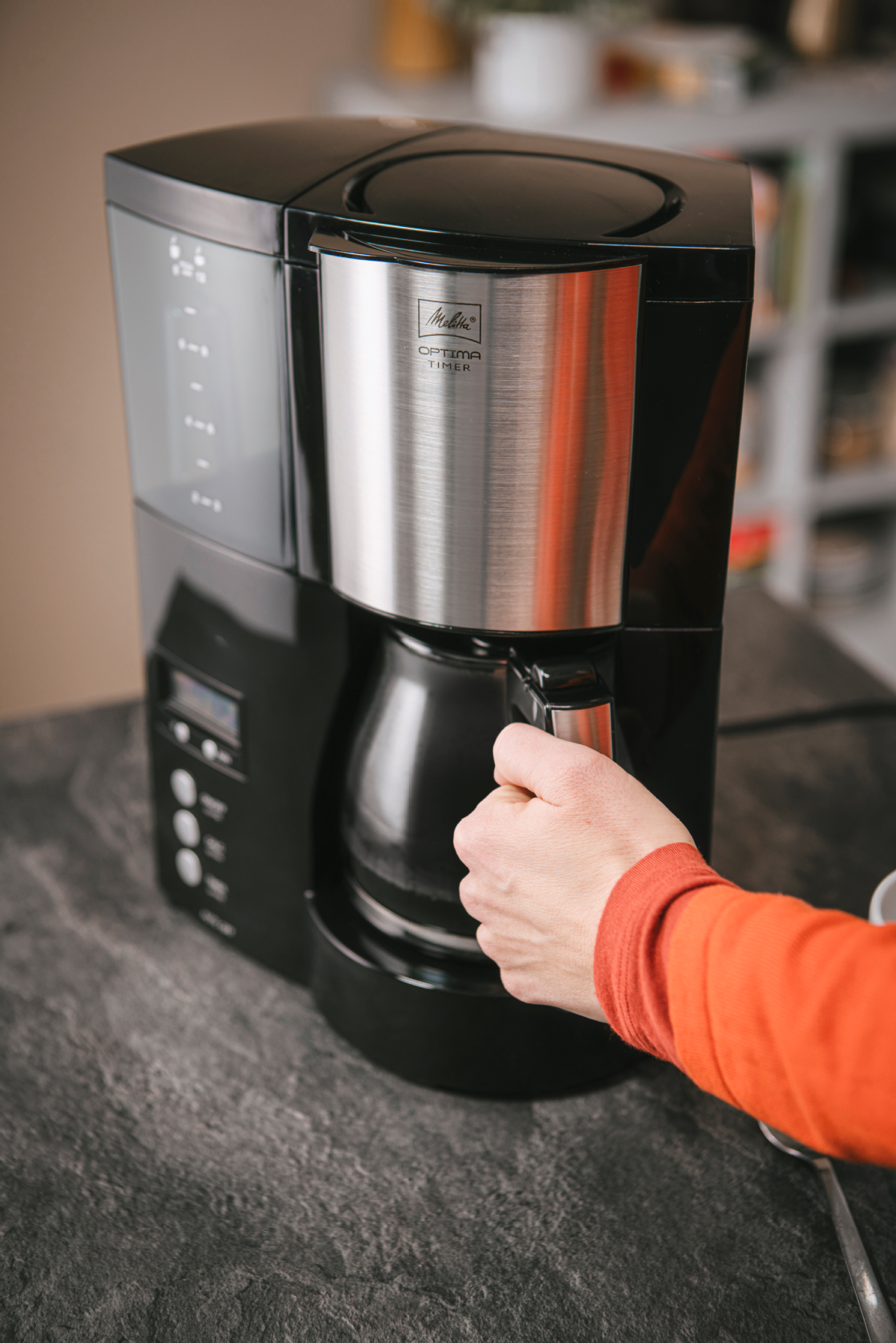 Melitta Optima® Timer Filterkaffeemaschine mit Glaskanne auf einer Küchenarbeitsplatte, mit einer Hand an der Kanne.