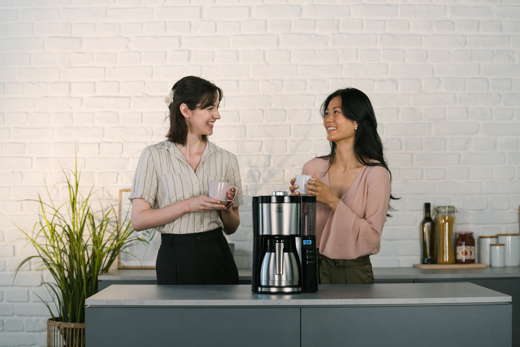 Melitta® LOOK® Therm Timer Filterkaffeemaschine mit Isolierkanne auf einer Küchenarbeitsplatte, mit zwei Personen und Tassen im Hintergrund.
