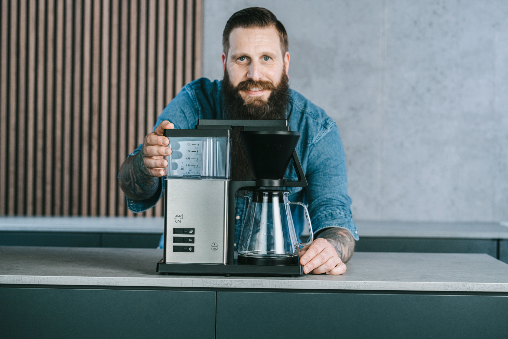 Filterkaffeemaschine mit Glaskanne auf einer Küchenarbeitsplatte, mit einer Person im Hintergrund.