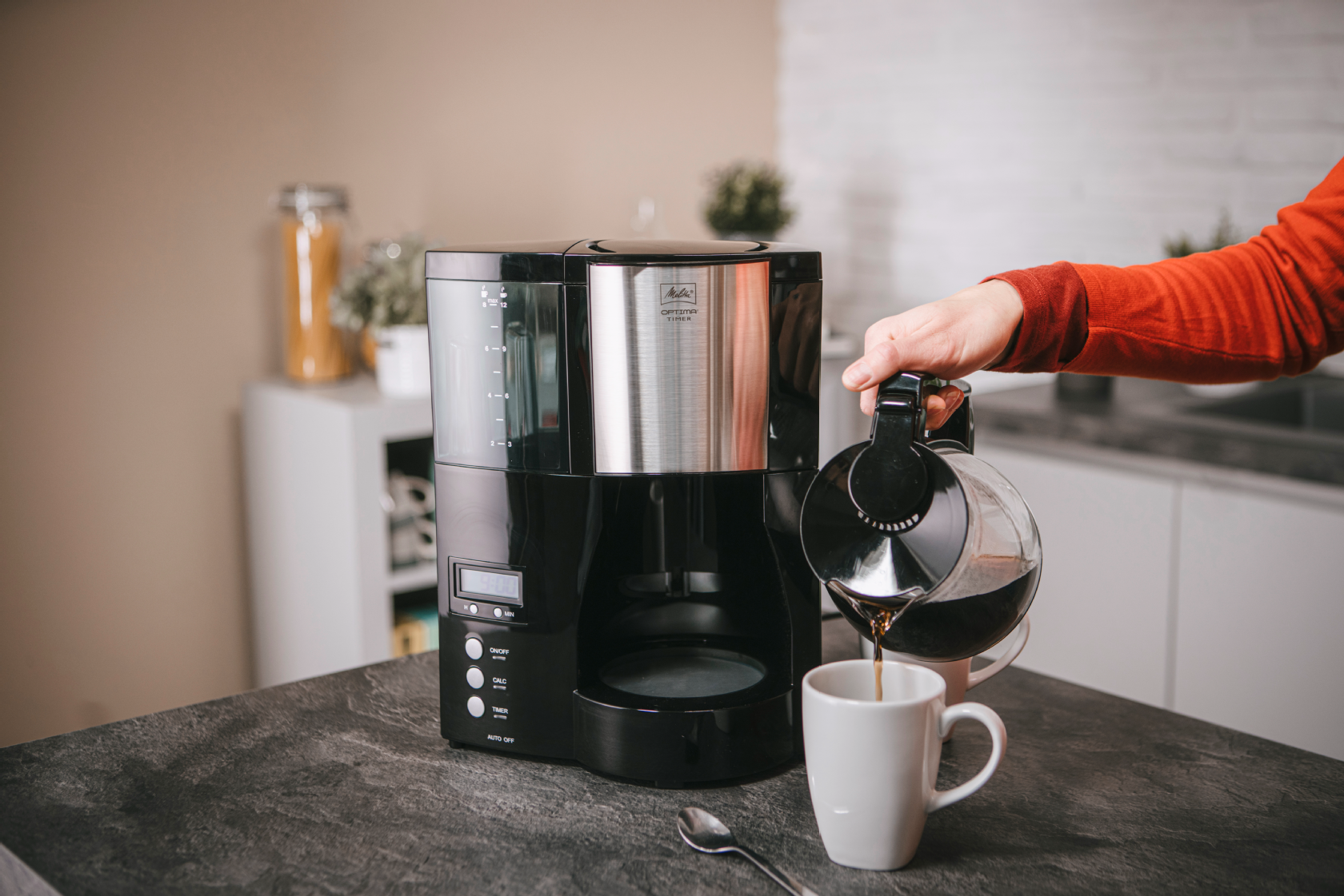 Melitta Optima® Timer Filterkaffeemaschine mit Glaskanne auf einer Küchenarbeitsplatte, während Kaffee in einen Becher gegossen wird.