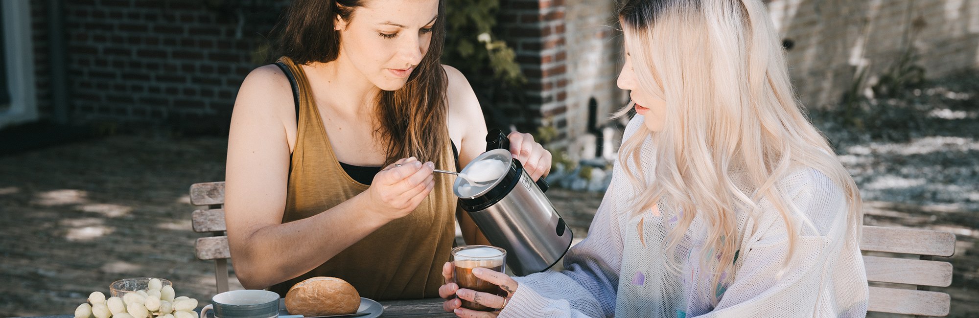 2 Frauen sitzen am Frühstückstisch und nehmen Milchschaum aus dem Cremio® Milchaufschäumer.