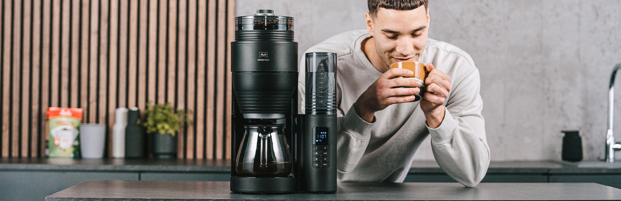 Melitta® AromaFresh X Filterkaffeemaschine mit Isolierkanne auf einer Küchenarbeitsplatte, mit einer Person und Tasse im Hintergrund