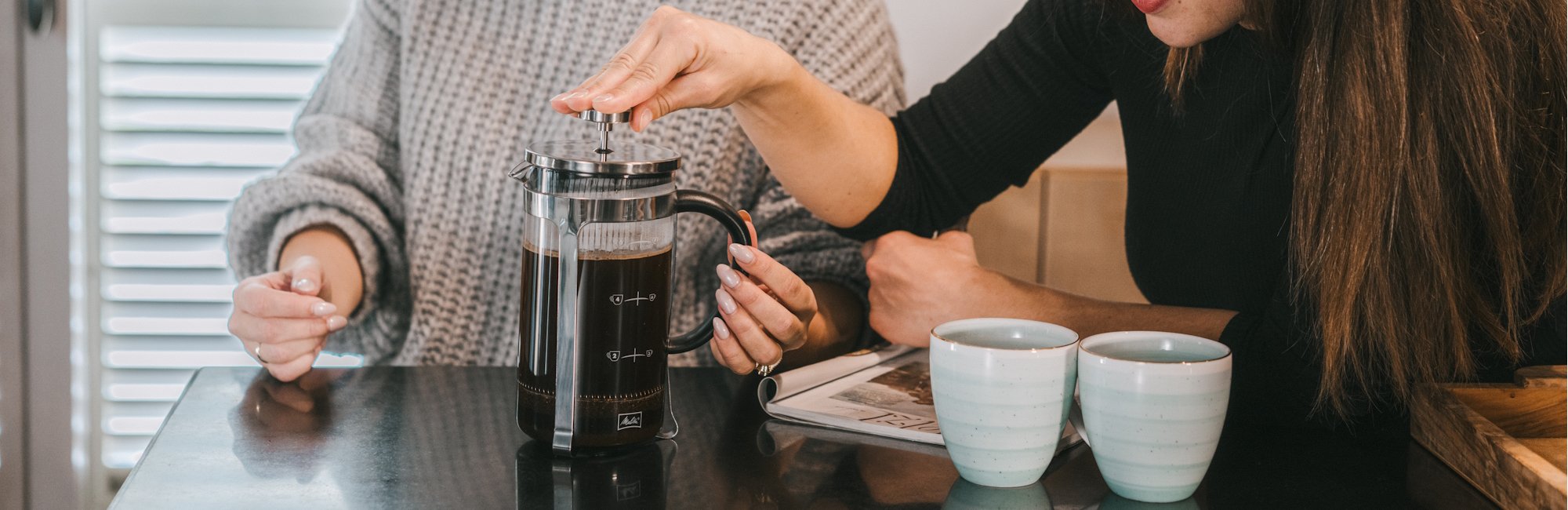 Frau drückt mit der Hand Kaffee in der FrenchPress herunter, daneben stehen zwei Tassen