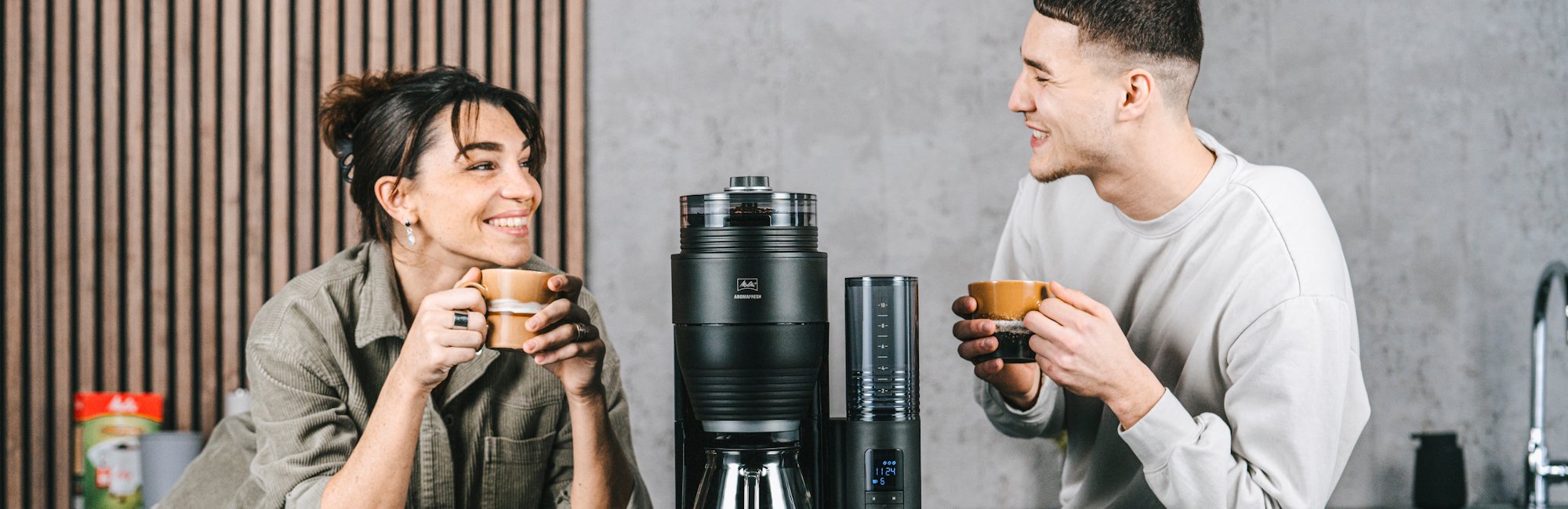 Melitta® AromaFresh X Filterkaffeemaschine mit Glaskanne und abnehmbarem Wassertank auf einer Küchenarbeitsplatte, mit zwei Personen und Tassen