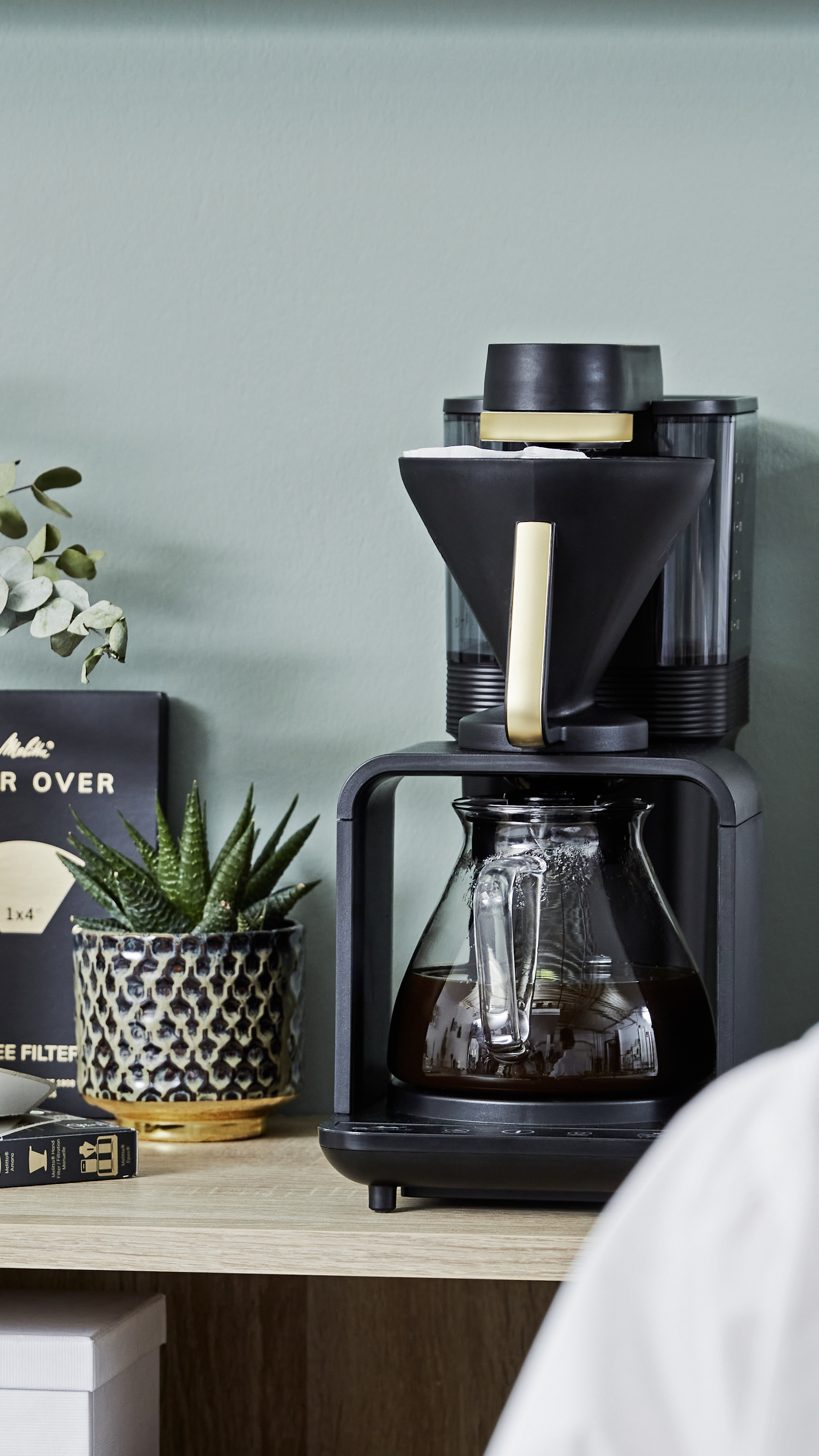Melitta epour Filterkaffeemaschine in Schwarz mit gefüllter Glaskanne auf einem Regal neben einer Pflanze
