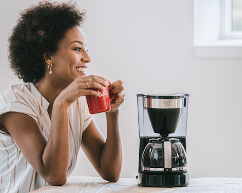 Lächelnde Frau mit roter Melitta-Tasse neben einer Melitta Filterkaffeemaschine mit Glaskanne