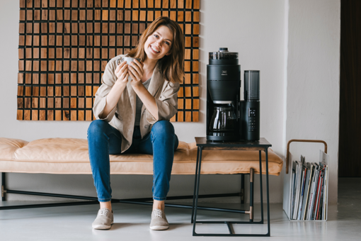 Lächelnde Frau mit Kaffeetasse sitzt auf einer Bank neben einer Melitta AromaFresh Filterkaffeemaschine