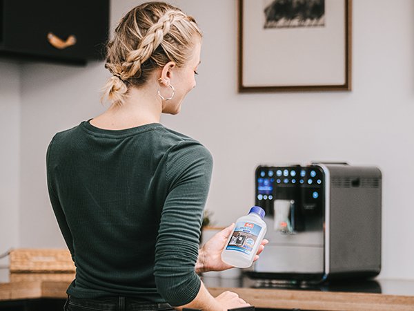Frau mit Melitta® Maschinenpflege in der Hand steht vor einem Barista Kaffeevollautomaten