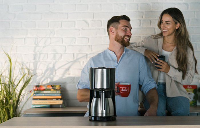 Melitta Enjoy Top Therm Filterkaffeemaschine vor lächelndem Paar mit Kaffeetassen.
