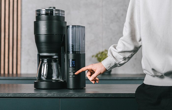 Melitta® AromaFresh X Filterkaffeemaschine mit Glaskanne und abnehmbarem Wassertank auf einer Küchenarbeitsplatte, mit einer Hand am Bedienfeld