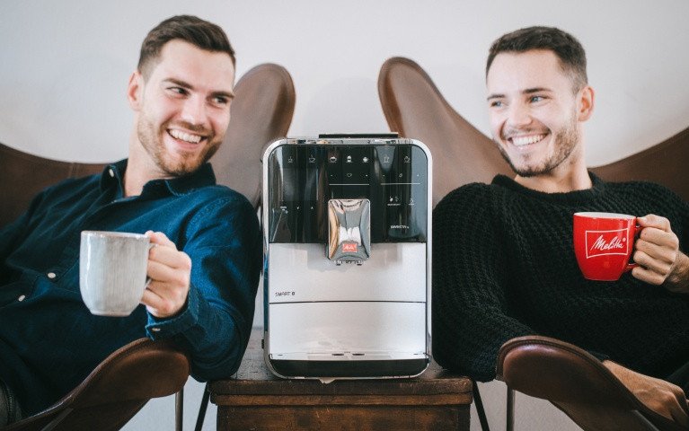 Zwei Männer genießen Kaffee mit einer silbernen Melitta CI Touch zwischen sich